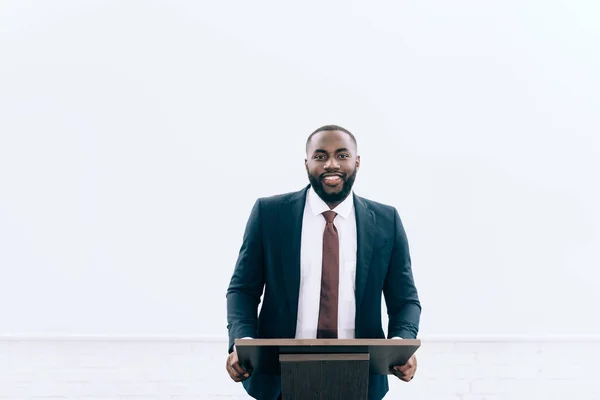 Sourire beau conférencier afro-américain debout sur le podium tribune pendant le séminaire dans la salle de conférence et en regardant la caméra — Photo de stock