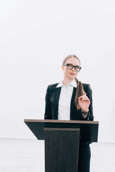 Beautiful lecturer standing at podium tribune and gesturing during seminar in conference hall — Stock Photo
