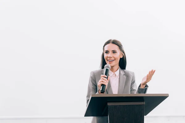 Attractive smiling lecturer talking into microphone and gesturing at podium tribune during seminar in conference hall — Stock Photo