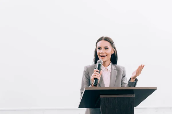 Attraente docente sorridente parlando in microfono e gesticolando al tribuna podio durante il seminario in sala conferenze — Foto stock