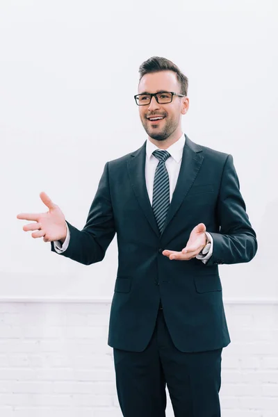 Beau conférencier souriant dans des lunettes parlant et gestualisant pendant le séminaire dans la salle de conférence — Photo de stock