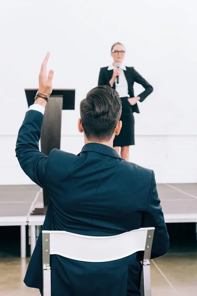 Atractivo orador hablando en el micrófono durante el seminario en la sala de conferencias, participante levantando la mano - foto de stock