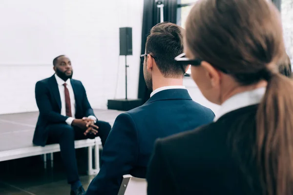 Enfoque selectivo del orador afroamericano sentado en el escenario durante el seminario en la sala de conferencias - foto de stock
