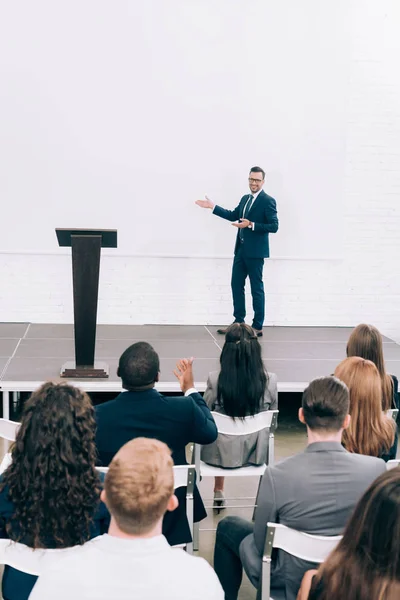 Smiling lecturer talking to multicultural audience during seminar and pointing on screen in conference hall — Stock Photo