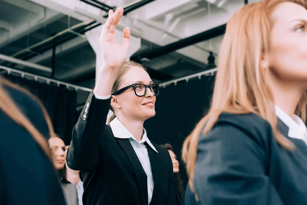 Donna d'affari sorridente negli occhiali con la mano in su desidera fare la domanda durante il seminario di affari nella sala conferenze — Foto stock