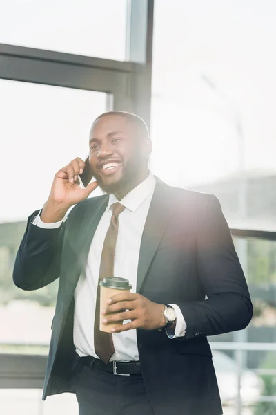 Sonriente hombre de negocios afroamericano con café para ir a hablar en smartphone en la sala de conferencias - foto de stock
