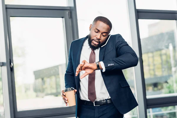 Empresário afro-americano com café para ir verificar o tempo enquanto conversa no smartphone na sala de conferências — Fotografia de Stock