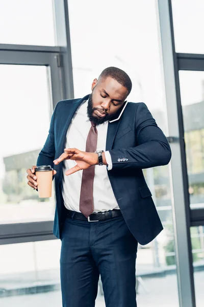 Hombre de negocios afroamericano con café para ir revisando el tiempo mientras mantiene una conversación en el teléfono inteligente en la sala de conferencias - foto de stock