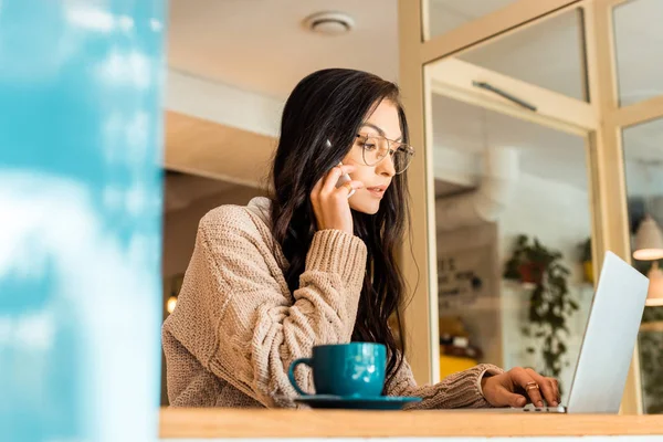 Belle femme assise dans un café avec ordinateur portable et parlant par smartphone — Photo de stock