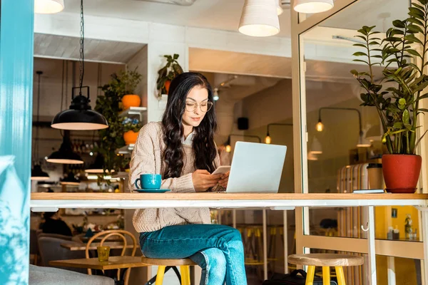 Schöne Frau sitzt mit Laptop im Café und benutzt Smartphone — Stockfoto