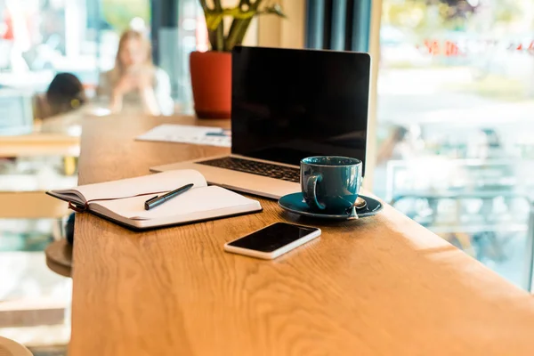 Laptop and smartphone with blank screens, notebook and cup of tea on wooden cafe counter — Stock Photo