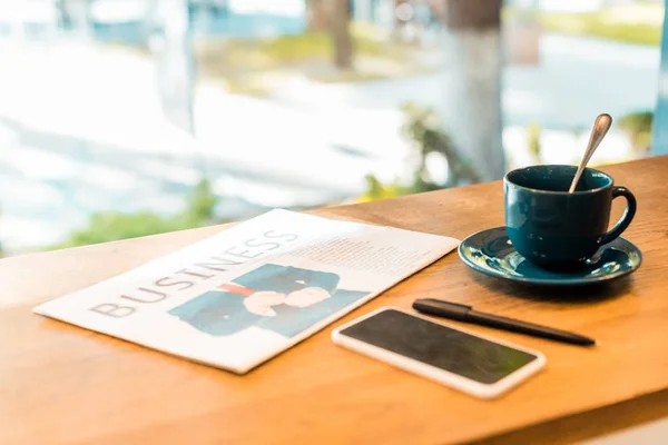 Teléfono inteligente con pantalla en blanco, periódico de negocios y taza de café en la mesa en la cafetería - foto de stock