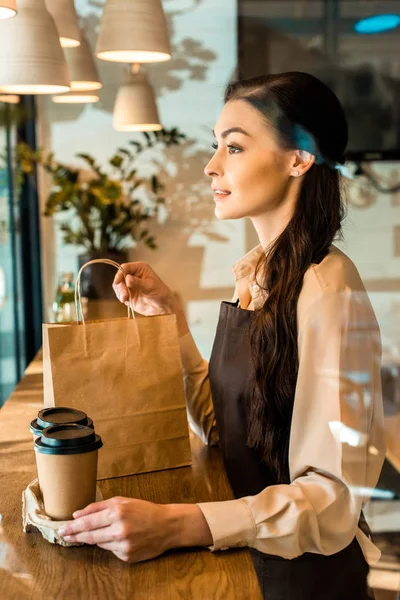 Vista lateral de la hermosa camarera en delantal con tazas de café desechables y bolsa de papel en la cafetería - foto de stock