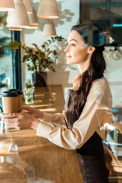 Vista lateral de la hermosa camarera en delantal sosteniendo tazas de café desechables en la cafetería - foto de stock