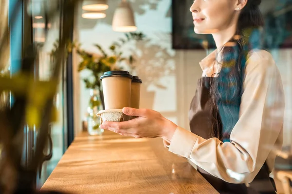 Обрезанное изображение официантки в фартуке, держащей кофе в бумажных чашках в кафе — стоковое фото
