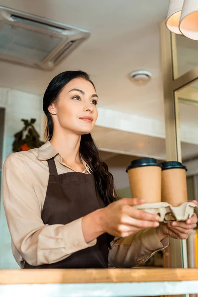 Vista de ángulo bajo de camarera morena sosteniendo café en tazas de papel en la cafetería - foto de stock