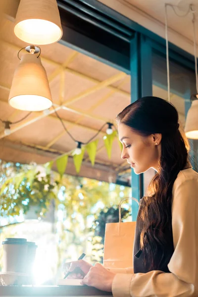 Vista lateral de camarera atractiva en delantal escribiendo algo a cuaderno en la cafetería - foto de stock