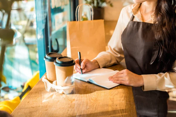 Imagen recortada de camarera en delantal escribiendo algo a cuaderno en la cafetería - foto de stock
