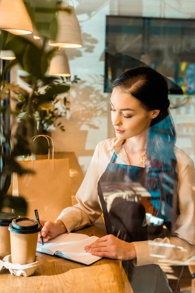Blick durch das Glas einer attraktiven Kellnerin in Schürze, die im Café etwas zum Notizbuch schreibt — Stockfoto