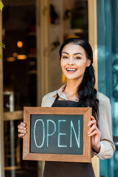 Happy beautiful cafe owner in apron holding open signboard near cafe and looking at camera — Stock Photo