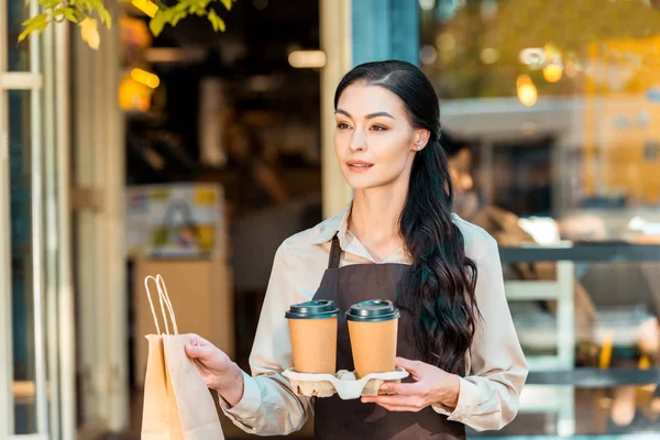 Attrayant serveuse dans tablier tenant deux café pour aller et sac en papier près du café — Photo de stock
