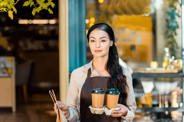 Привлекательная официантка в фартуке с двумя одноразовыми чашками кофе и бумажным пакетом рядом с кафе — стоковое фото