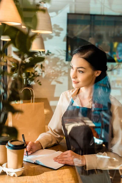 View through glass of beautiful waitress in apron writing something to notebook in cafe — Stock Photo