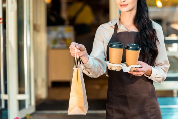 Cropped image of waitress in apron holding two coffee in paper cups and paper bag near cafe — Stock Photo