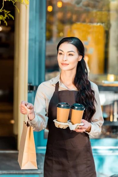 Hermosa camarera en delantal sosteniendo dos café en tazas de papel y bolsa de papel cerca de la cafetería - foto de stock