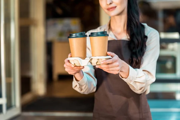 Image recadrée de serveuse dans tablier tenant deux café dans des tasses en papier près du café — Photo de stock