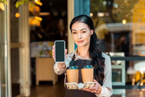 Schöne Kellnerin in Schürze hält Coffee to go und zeigt Smartphone mit leerem Bildschirm in der Nähe des Cafés — Stockfoto
