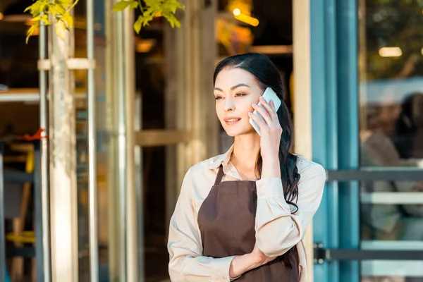 Schöne Kellnerin in Schürze, die mit dem Smartphone spricht und in der Nähe eines Cafés wegschaut — Stockfoto