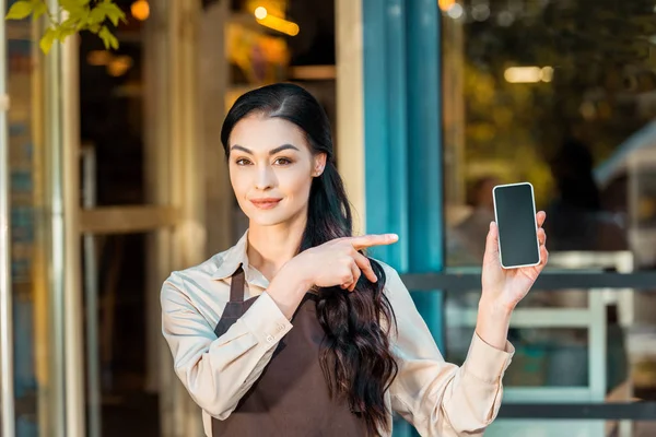 Красива офіціантка в фартусі, що вказує на смартфон з порожнім екраном біля кафе — стокове фото
