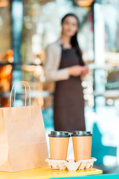 Waitress in apron standing near cafe with coffee in paper cups and paper bag on foreground — Stock Photo