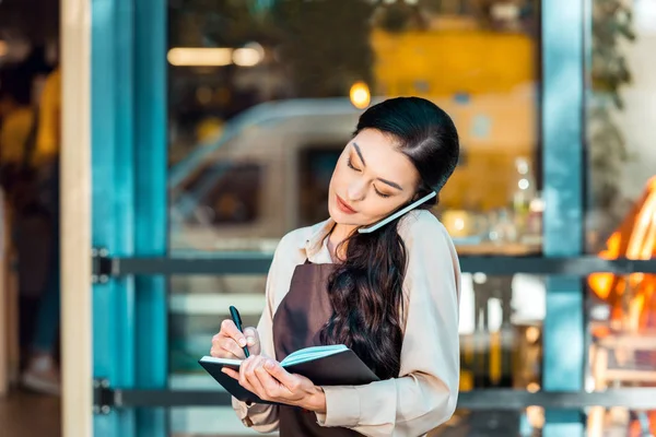 Attraktive Kellnerin, die auf der Straße in der Nähe eines Cafés mit dem Smartphone spricht und Notizen zum Notizbuch macht — Stockfoto