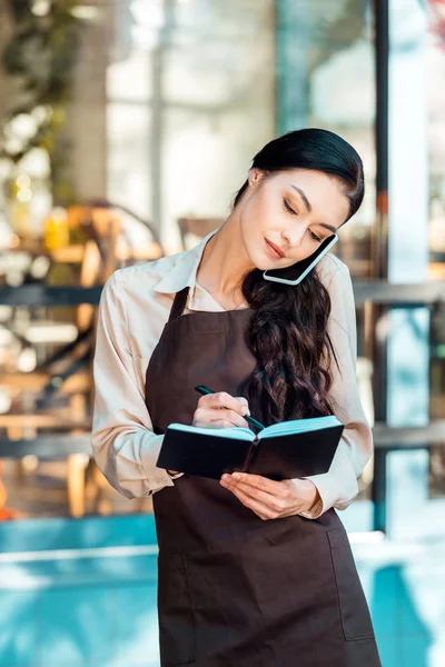 Beautiful waitress in apron talking by smartphone on street near cafe and taking notes to notebook — Stock Photo