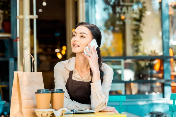 Belle serveuse dans tablier assis à table et parlant par smartphone dans la rue près du café — Photo de stock