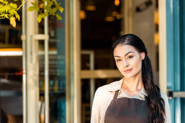 Retrato de hermosa camarera en delantal mirando la cámara cerca de la cafetería — Stock Photo