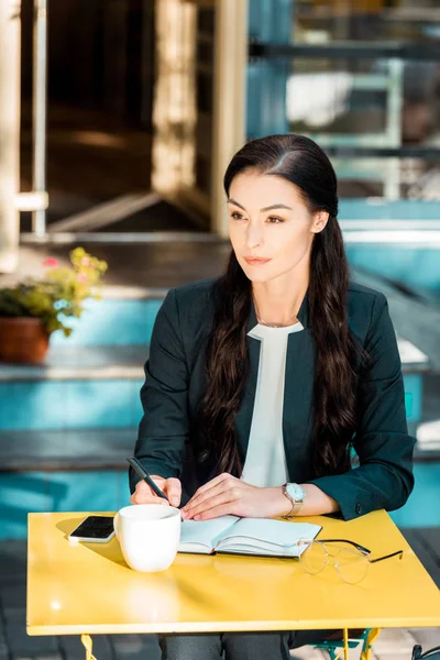 Schöne Geschäftsfrau schreibt im Straßencafé etwas ins Notizbuch und schaut weg — Stockfoto