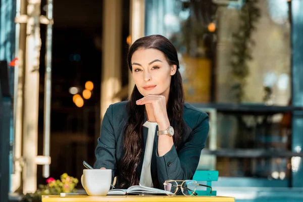 Hermosa mujer de negocios descansando barbilla en la mano y mirando a la cámara en la cafetería de la calle - foto de stock