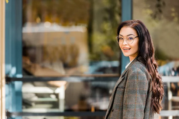 Schöne lächelnde Frau mit Brille, die auf der Straße in der Nähe eines Cafés in die Kamera schaut — Stockfoto
