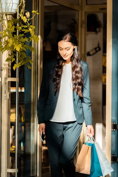 Hermosa mujer caminando desde la tienda con bolsas de compras y la celebración de teléfono inteligente - foto de stock