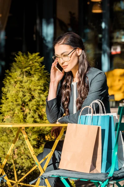 Attractive brunette woman talking by smartphone at table at street cafe, shopping bags on chair — Stock Photo
