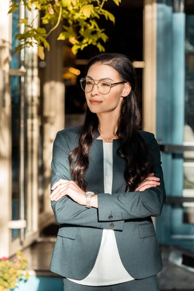 Beautiful cafe owner in glasses standing with crossed arms on street near cafe and looking away — Stock Photo