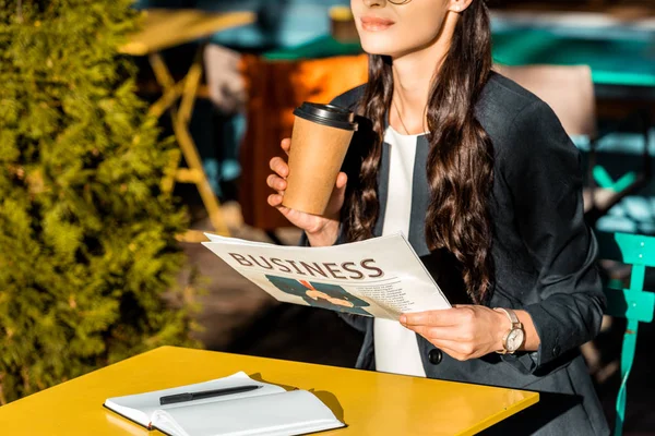 Cropped view of woman with coffee to go, business newspaper and planner sitting on cafe terrace — Stock Photo