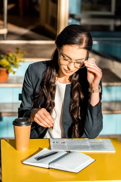 Концентрована жінка читає газету з кавою, щоб піти і планувальник на терасі кафе — стокове фото