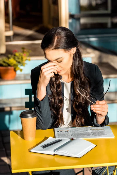 Mulher cansada com café para ir, jornal e planejador segurando óculos e sentado no terraço do café — Fotografia de Stock