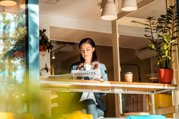 Brünette Geschäftsfrau liest Wirtschaftszeitung im Café mit Laptop und Kaffeetasse — Stockfoto