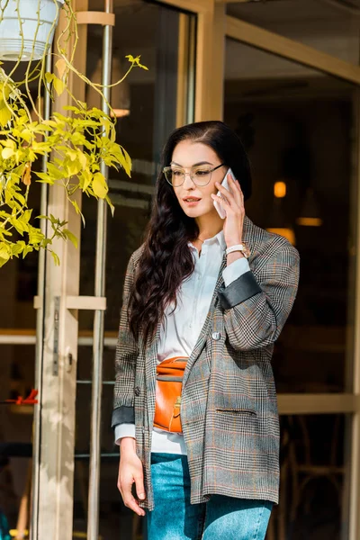 Atractiva mujer en chaqueta de moda hablando en el teléfono inteligente cerca de la entrada del restaurante - foto de stock