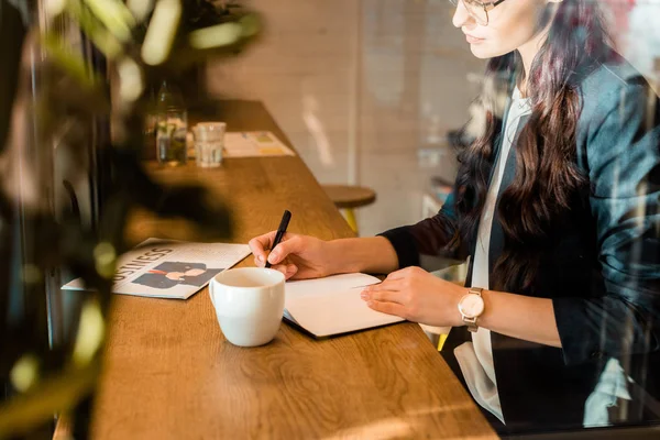 Freelancer feminino escrevendo em diário à mesa com xícara de café e jornal de negócios no café — Fotografia de Stock